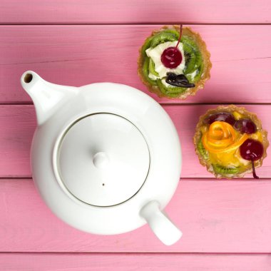 Kivi ile yapılan renkli meyve kekler, turuncu, şekerlenmiş kiraz, krema ve çikolata ile bir çaydanlık pembe bir arka plan üzerinde. Yukarıdan görüntülemek.