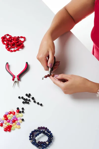 Jewelry making. Making bracelet of colorful beads. Female hands with a tool on a white background.