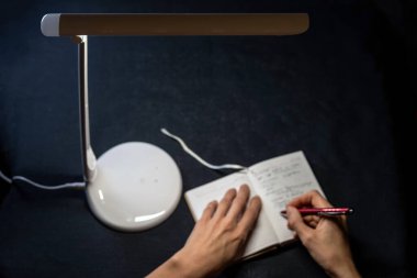 Koyu mavi arka planda bir masa lambasının ışığında elinde kalem ve not defteriyle. Seçici odak.  