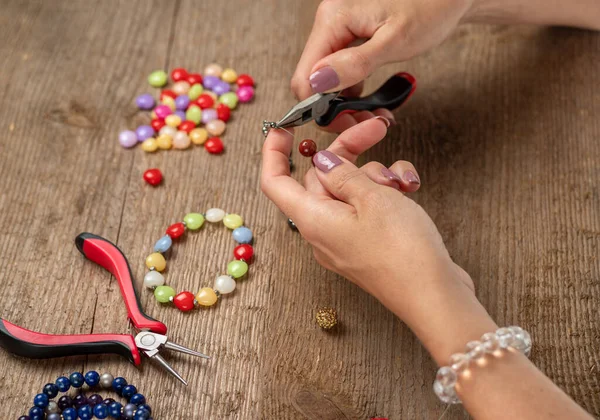 Jewelry making. Making a bracelet of colorful beads. Female hands with a tool on a rough wooden table. Selective focus