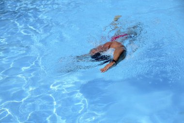 Kafkasyalı bir adam güneşli bir günde açık havuzda serbest yüzer. Adam kırmızı bir mayo ve siyah bir şapka giyiyor. Boş zamanlarında yüzen zinde bir adam.