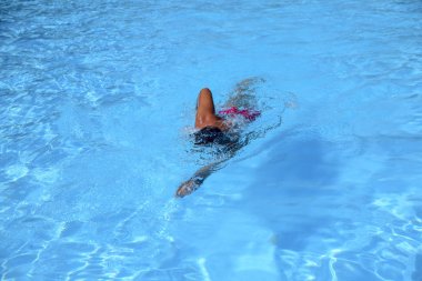 Kafkasyalı bir adam güneşli bir günde açık havuzda serbest yüzer. Adam kırmızı bir mayo ve siyah bir şapka giyiyor. Boş zamanlarında yüzen zinde bir adam.