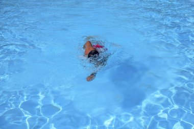 Kafkasyalı bir adam güneşli bir günde açık havuzda serbest yüzer. Adam kırmızı bir mayo ve siyah bir şapka giyiyor. Boş zamanlarında yüzen zinde bir adam.