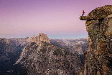 A fearless male hiker is standing on an overhanging rock at Glacier Point enjoying the breathtaking view towards famous Half Dome in beautiful post sunset twilight in summer, Yosemite National Park, California clipart