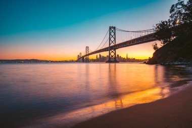 Ünlü Oakland Bay Köprüsü 'nün klasik panoramik manzarası. San Francisco' nun ufuk çizgisi güzel bir alacakaranlıkla aydınlanıyor. Yaz aylarında günbatımı parlıyor.