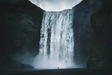 Sarı yağmurluklu yürüyüşçünün karamsar bir günde dev bir Skogafoss şelalesinin önünde duruşu yaz mevsiminde kara bulutlar, Skogar, İzlanda