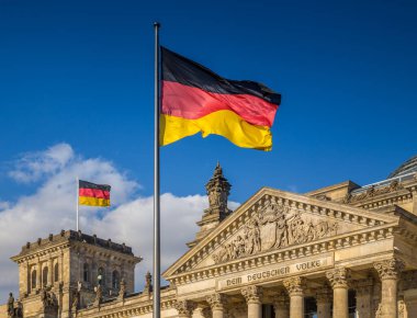 Alman bayrakları, Alman Parlamentosu 'nun (Deutscher Bundestag) ünlü binasında, mavi gökyüzü ve bulutlarla güneşli bir günde, Almanya' nın orta Berlin Mitte bölgesinde rüzgarda sallanıyor.