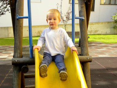 Yakışıklı üç yaşındaki çocuk sarı bir plastik kaydırağın üzerinde oturuyor ve onu terk etmek üzere.        