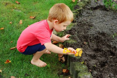 Sarışın bir çocuk yeşil çimenlerde oturur ve çiçek tarhındaki toprağı gevşetir. Üç yaşında bir çocuk elinde oyuncak bir spatulayla toprağı kazıyor..