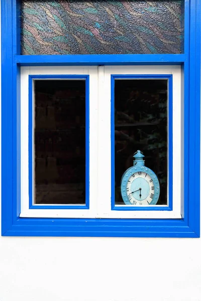 Old clock painted in blue-inner ledge of a white and blue painted wooden casement window-stained glass upper section housed in the white wall of a building-street in old central Kas town. Lycia-Turkey