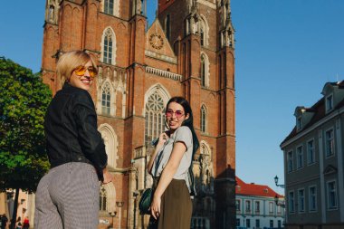 two attractive smiling women standing in front of wroclaw cathedral and looking at camera clipart
