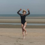 Beautiful young woman in black bodysuit jumping on shore on cloudy day