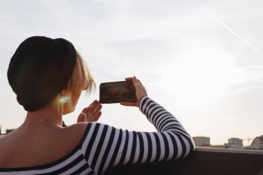 çizgili gömlekli fotoğrafta smartphone ile günbatımı gökyüzü önünde genç kadının arka görünüm