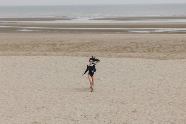 attractive young woman in black bodysuit running on sandy seashore clipart