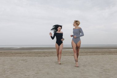 happy young women in bodysuits running by sandy beach on cloudy day clipart