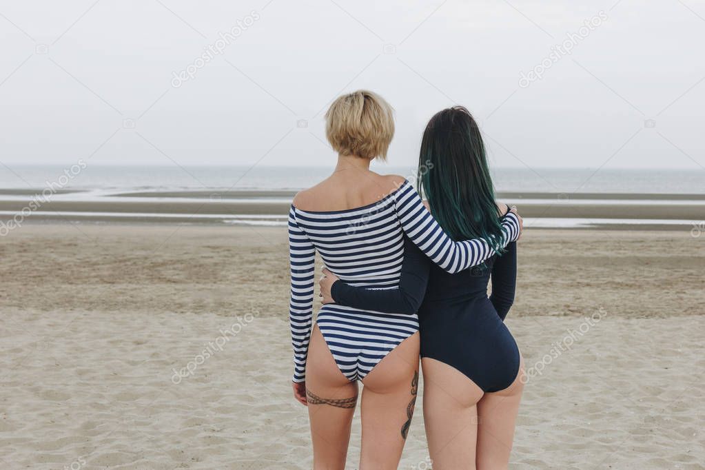 rear view of sexy young women in bodysuits embracing on sandy beach on cloudy day