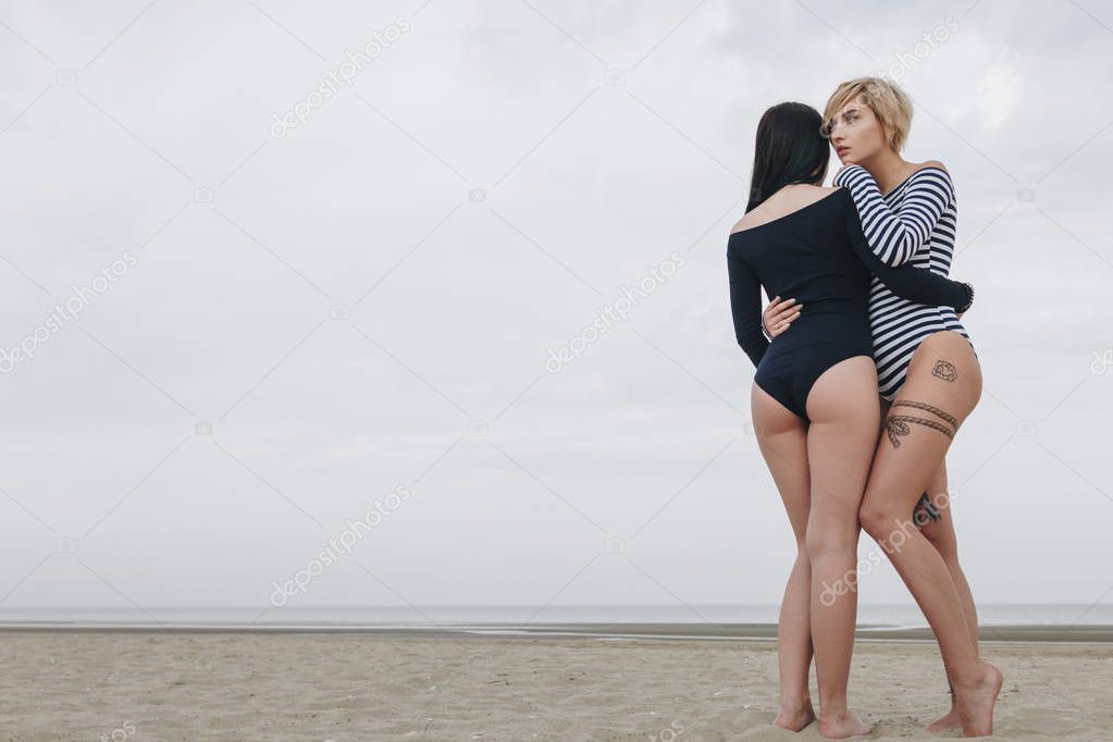 rear view of young women in bodysuits embracing on sandy beach on cloudy day