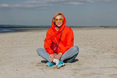 beautiful smiling girl in red hoodie sitting on beach, saint michaels mount, France clipart