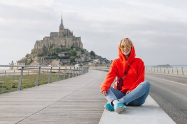 happy young woman smiling at camera while sitting near mont saint michel, france  clipart