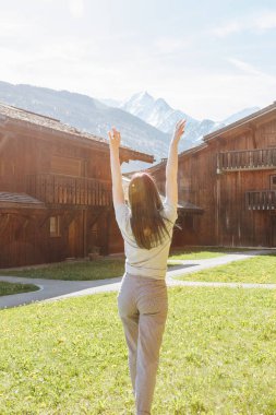 back view of beautiful young woman raising hands while standing between wooden houses in mountain village, mont blanc, alps clipart