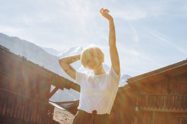 beautiful happy young woman raising hand and looking at camera while standing between wooden houses in mountain village, mont blanc, alps clipart