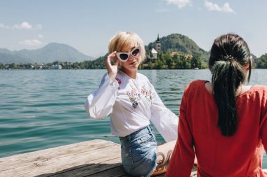 güzel kız kameraya dağ Gölü, bled, ahşap iskele üzerinde sevgilisiyle otururken Slovenya seyir güneş gözlüğü