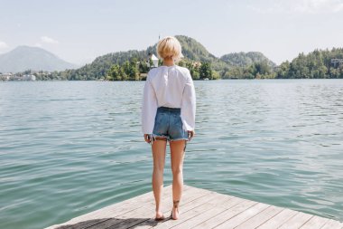 back view of young woman standing on wooden pier and looking at scenic mountain lake, bled, slovenia clipart