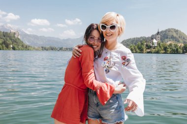 beautiful young women in sunglasses hugging and smiling at camera while standing near tranquil mountain lake, bled, slovenia clipart
