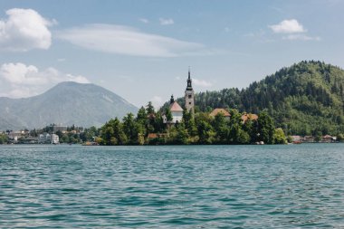 old architecture and green trees at bank on scenic mountain lake, bled, slovenia clipart