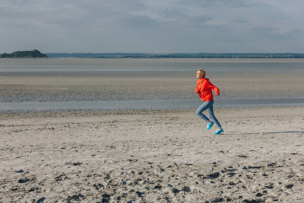 стильная молодая женщина, бегущая по песчаному пляжу, Сен-Мишель, Франция
