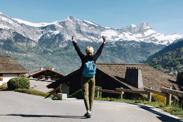 back view of girl with backpack raising hands and walking in scenic mountain village, mont blanc, alps