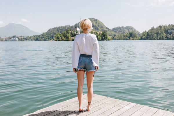 back view of young woman standing on wooden pier and looking at scenic mountain lake, bled, slovenia
