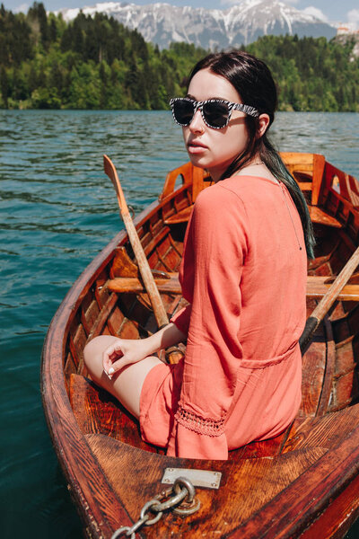 beautiful young woman in sunglasses sitting in boat at tranquil mountain lake, bled, slovenia