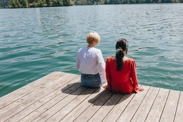 Back view of two young stylish women sitting on wooden pier at lake, bled, slovenia — Stock Image Back View Two Young Stylish Women Sitting Wooden Pier Lake Stock Picture