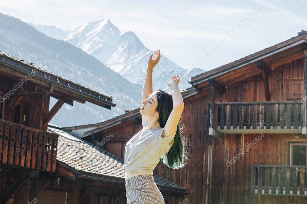 side view of beautiful young woman raising hands while standing between wooden houses in mountain village, mont blanc, alps