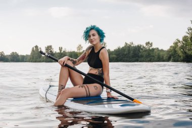 beautiful tattooed girl with blue hair sitting on paddleboard on river clipart