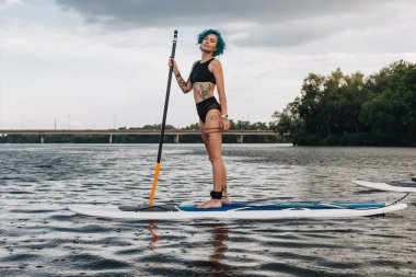 beautiful dreamy athletic tattooed woman standup paddleboarding on river clipart