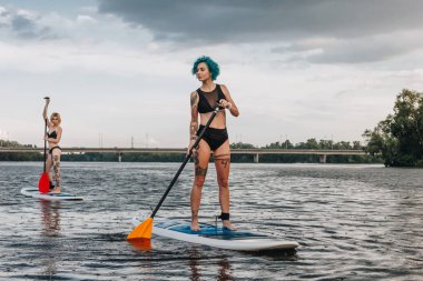 athletic women standup paddleboarding together on river clipart
