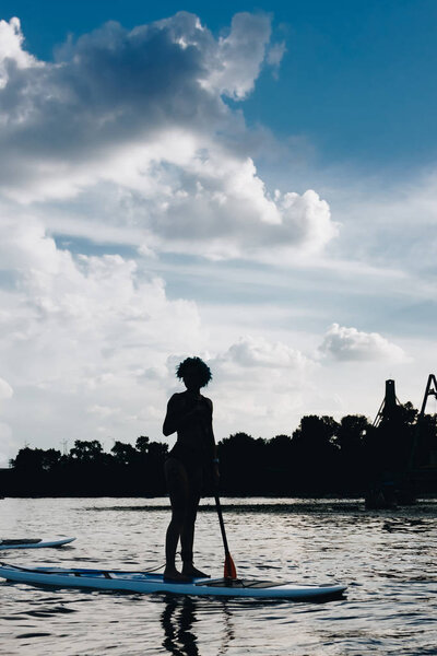 silhouette of sportswoman standing on paddle board on river with cloudy sky