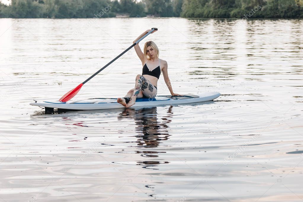 attractive tattooed sportswoman in bikini relaxing on paddle board on river