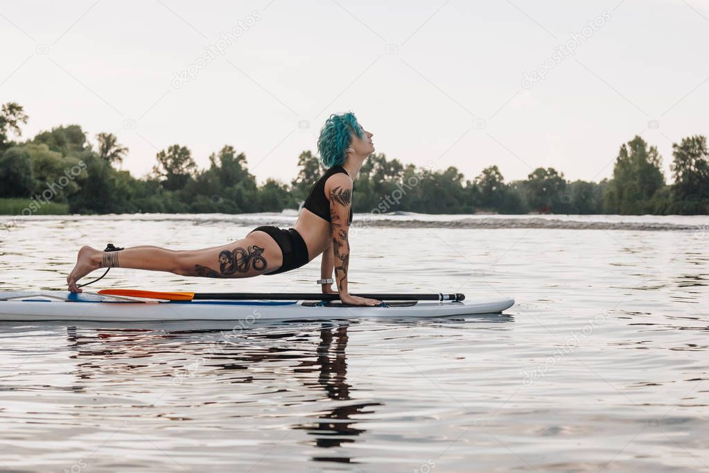 tattooed woman with blue hair practicing yoga on paddleboard in water. Upward facing dog pose (Urdhva Mukha Svanasana)