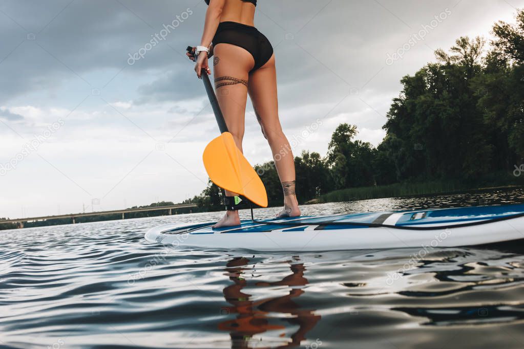 partial view of tattooed woman in bikini standing on sup board on river