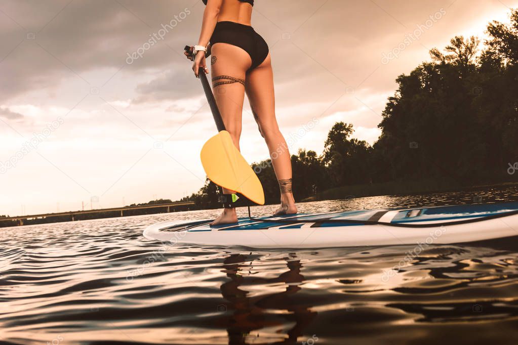 partial view of tattooed girl standing on sup board on river at sunset