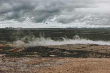 scenic view of landscape with volcanic vents under cloudy sky in Haukadalur valley in Iceland clipart