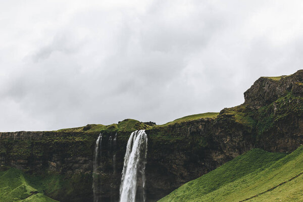 scenic view of landscape with Seljalandsfoss waterfall in highlands under cloudy sky in Iceland 