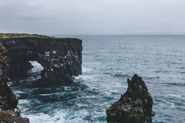 dramatic shot of rocky cliffs and stormy ocean in Arnarstapi, Iceland on cloudy day clipart