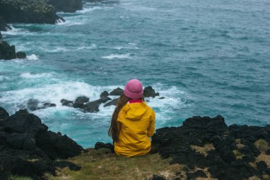 rear view of woman in yellow raincoat sitting on cliff in front of ocean, iceland clipart