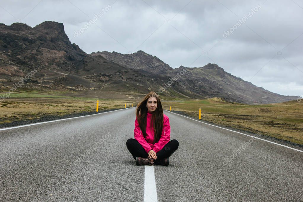Smiling young woman sitting on empty road in iceland with beautiful mountains on background