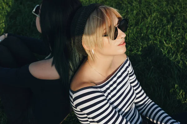 Mujeres jóvenes con estilo sentado en la hierba verde espalda con espalda - foto de stock