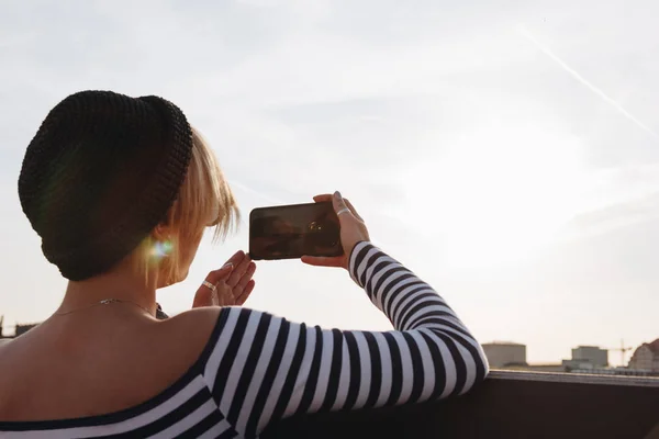 Vista trasera de la mujer joven en camisa a rayas tomando fotos con teléfono inteligente en frente del cielo del atardecer - foto de stock